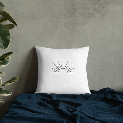 White pillow with sun design on a dark blue blanket against a gray wall.